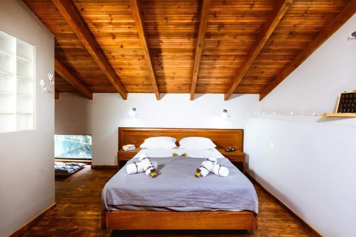 Une chambre avec un plafond en bois, un lit avec des oreillers blancs et une couverture grise, une table de chevet avec une lampe, et une fenêtre avec des stores.