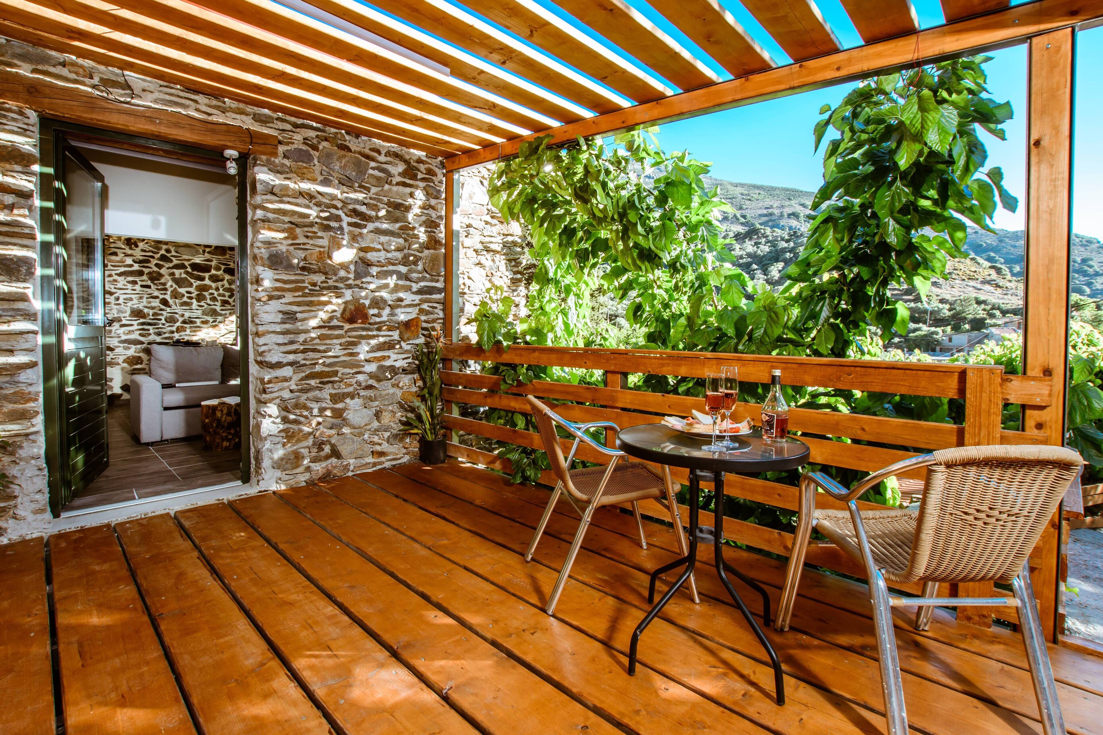 A wooden deck with a stone wall, a table with chairs, and a view of greenery and hills.