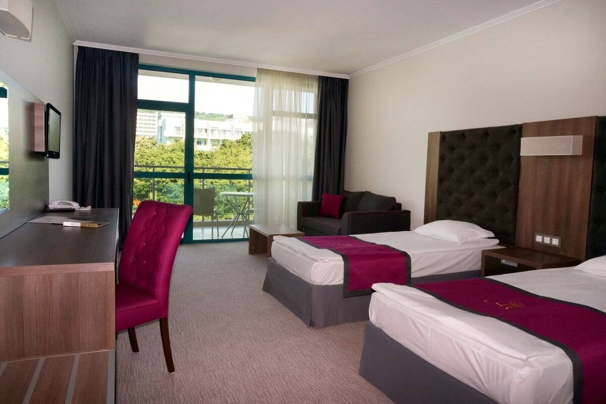 A hotel room with two beds, a desk, a chair, and a balcony with a view of greenery.
