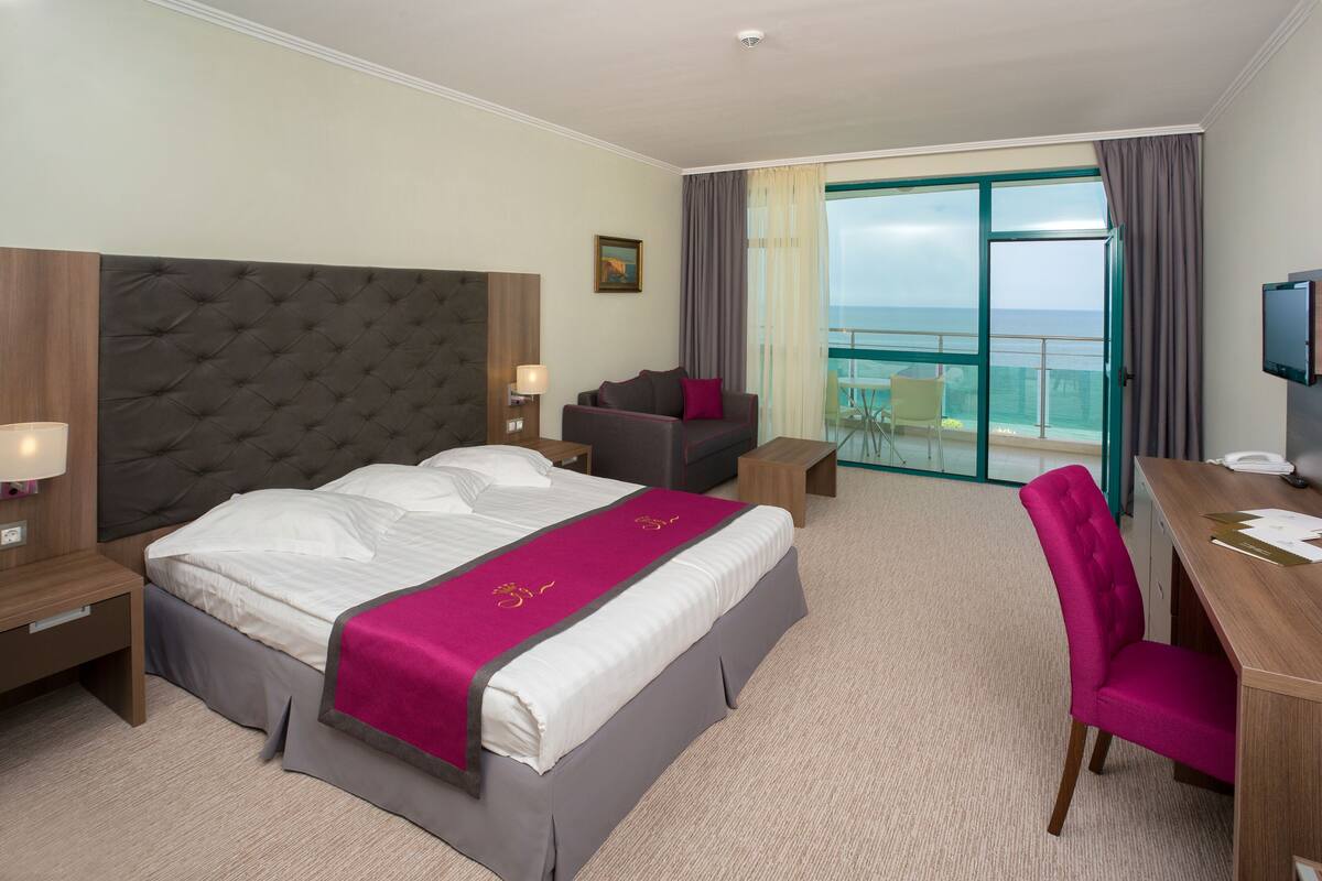 A hotel room with a large bed, a desk, a chair, a sofa, and a balcony with a view.