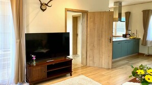 Una sala de estar con televisor sobre un mueble de madera, una zona de cocina con armarios y una ventana, y una puerta que da a otra habitación.
