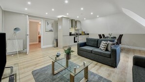 A modern living room with a glass coffee table, grey sofa, and a dining area with a kitchen in the background. - Deanway Serviced Apt Chalfont St Giles (Chalfont St Giles)