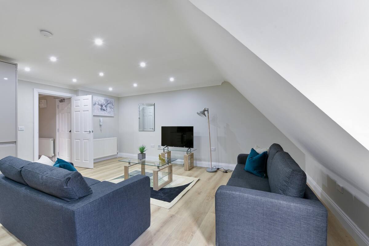 A modern living room with a loft-style ceiling, grey sofas, a glass coffee table, and a flat-screen TV.