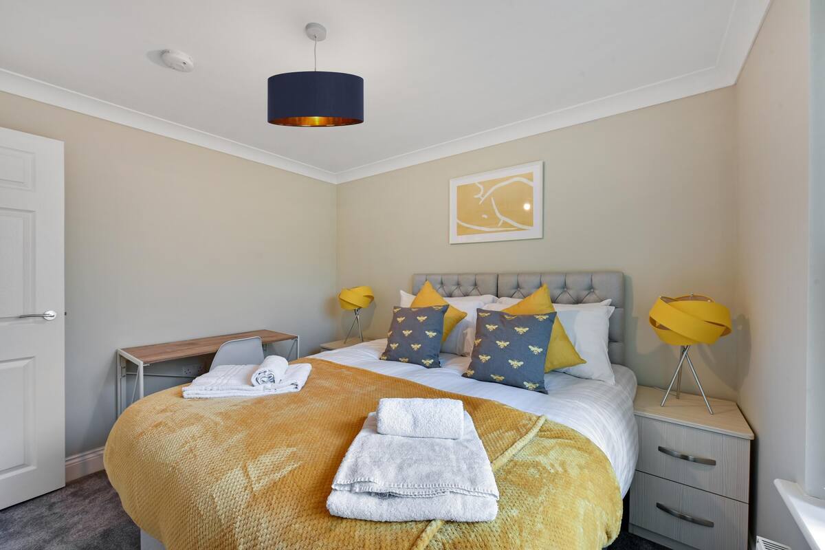 A neatly made bed with a yellow blanket and white towels, a bedside table with a lamp, and a framed picture on the wall.