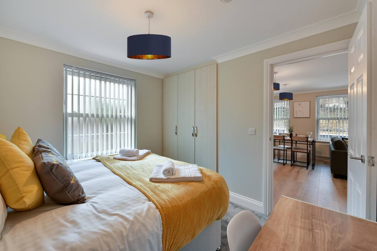 A bedroom with a bed, pillows, a bedside table, a window with blinds, and a door leading to a dining area.