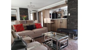 Relax in this stylish living room with a sofa, coffee table, and side table, featuring a chandelier and kitchen area with wooden cabinets.