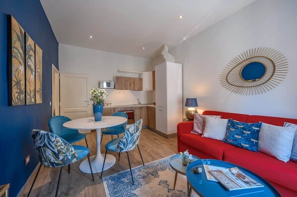 A modern living room with a red sofa, blue chairs, a round coffee table, and a kitchen in the background.