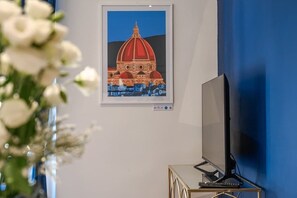 A framed artwork of a building, a television on a stand, and a vase with flowers.