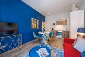 A modern living room with a blue accent wall, a red sofa, a dining area with a round table and chairs, a kitchen with wooden cabinets, and a flat-screen TV.