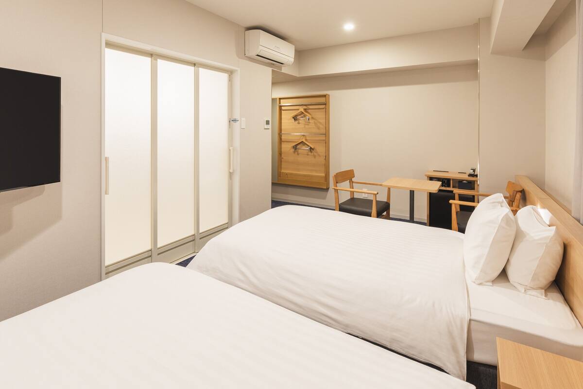 A hotel room with two beds, a desk, a chair, and a TV.
