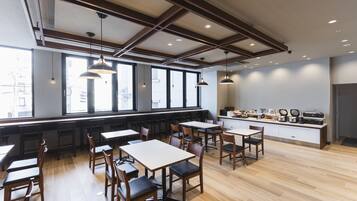 A modern restaurant with wooden tables and chairs, large windows, and a kitchen counter visible.