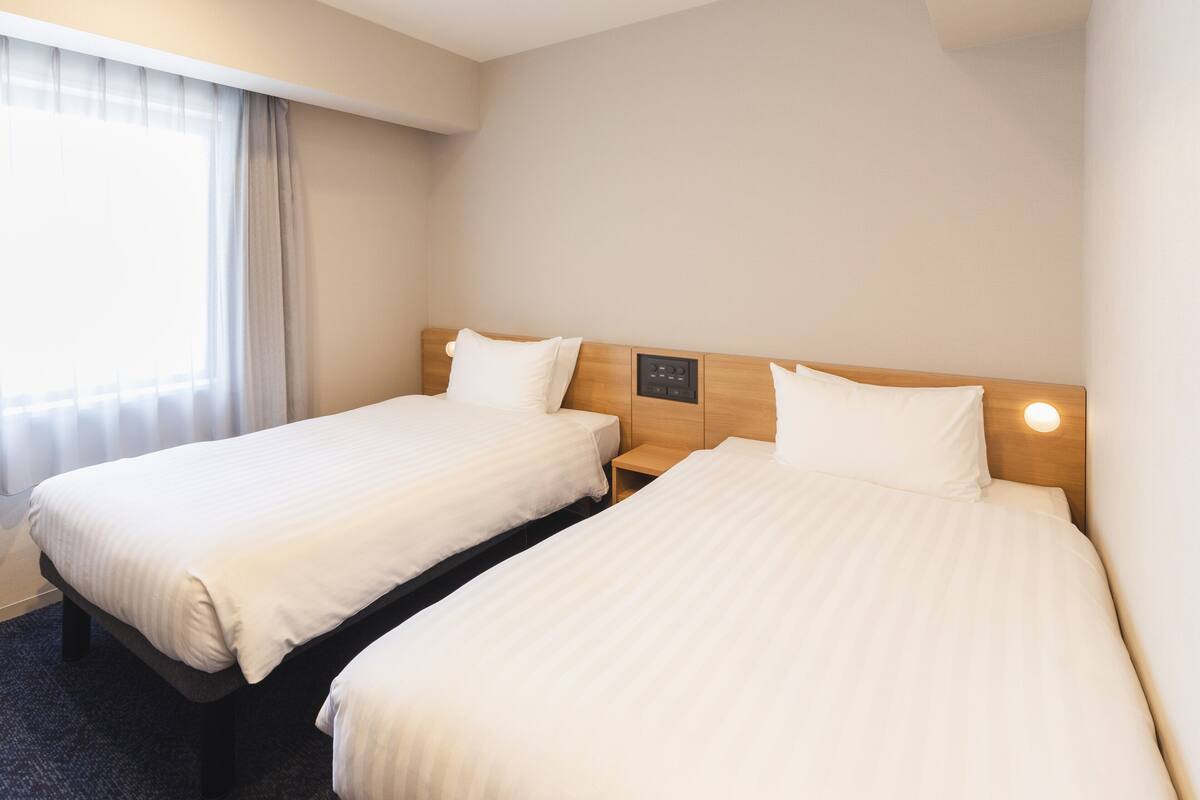 A hotel room with two single beds, a bedside table, a control panel, and a window with curtains.
