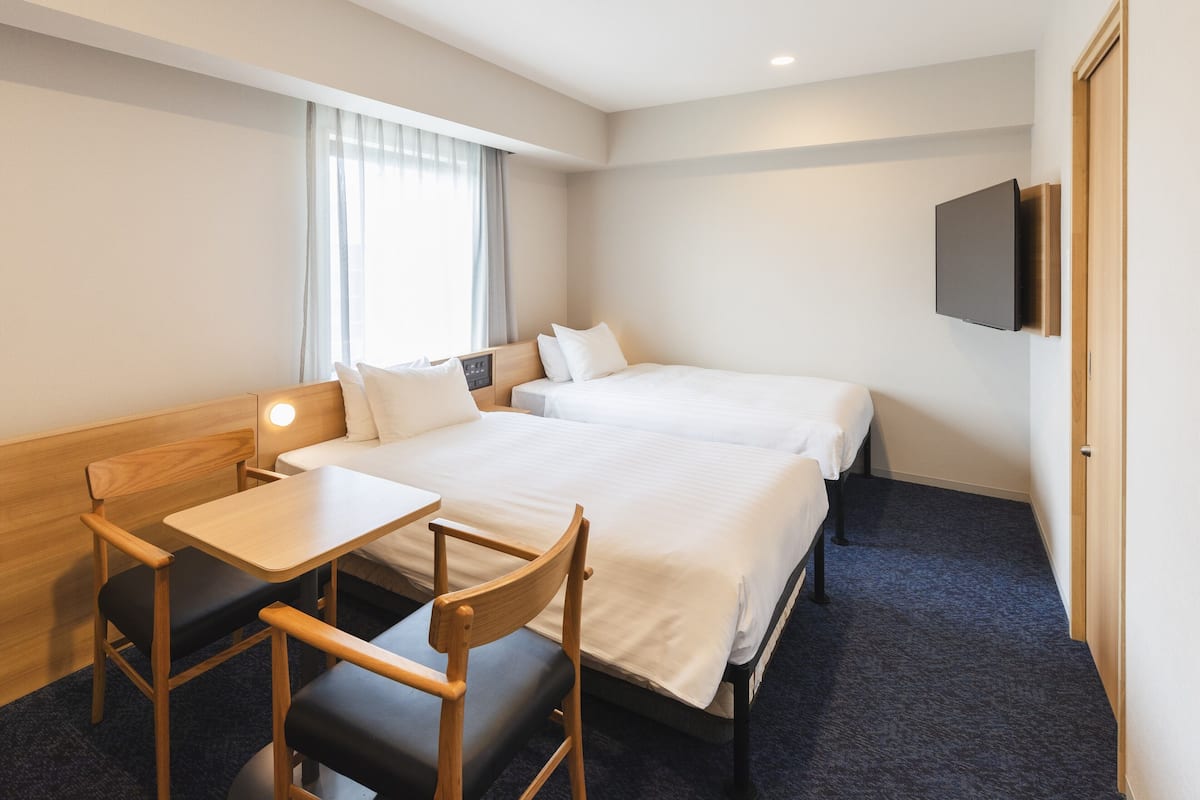 A hotel room with two beds, a desk, a chair, a TV mounted on the wall, and a window with curtains.