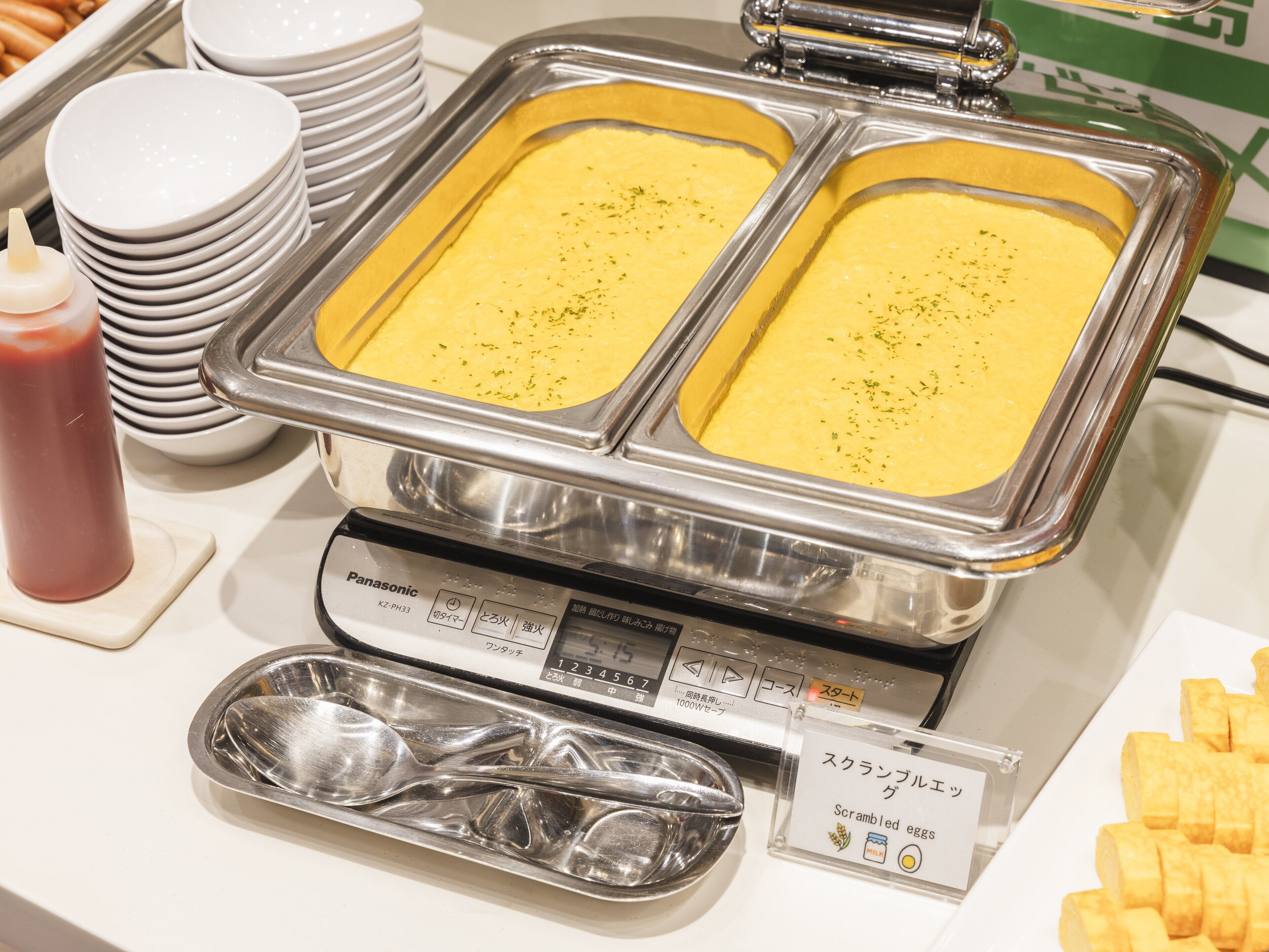 A buffet setup with scrambled eggs, ketchup, and a weighing scale.