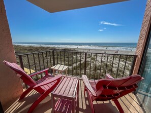 A balcony with red Adirondack chairs and a small table, overlooking a beach with sand dunes and the ocean.