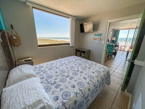 A bedroom with a bed, a window with a beach view, a TV on the wall, and a door leading to a balcony.