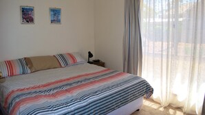A bedroom with a large bed, two framed pictures on the wall, a bedside table with a lamp, and a window with curtains.