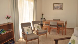 A living room with a wooden bookshelf, two armchairs with floral cushions, a dining table with four chairs, and a framed landscape painting on the wall.