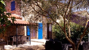 Un edificio de piedra con techo de tejas, mesas al aire libre y un árbol en primer plano.