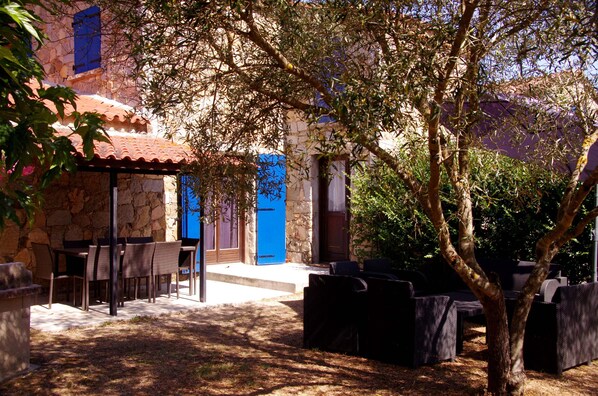 A stone building with a tiled roof, outdoor seating, and a tree in the foreground.