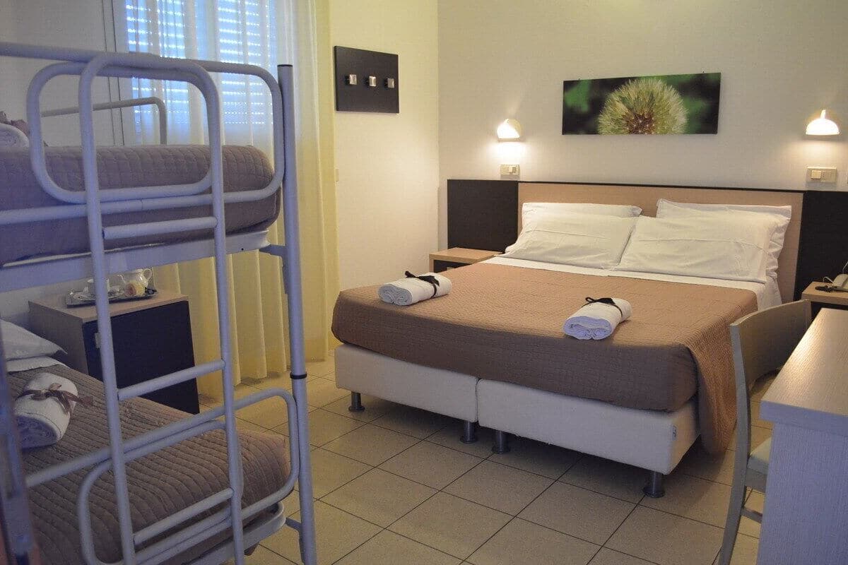 Une chambre d’hôtel avec des lits superposés, un lit simple avec une tête de lit, des tables de chevet, un bureau et une lampe fixée au mur.
