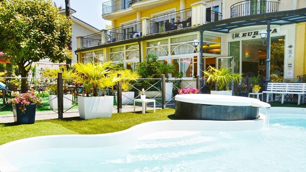 Una piscina con fuente, rodeada de un jardín bien cuidado con diversas plantas y un espacio para sentarse.