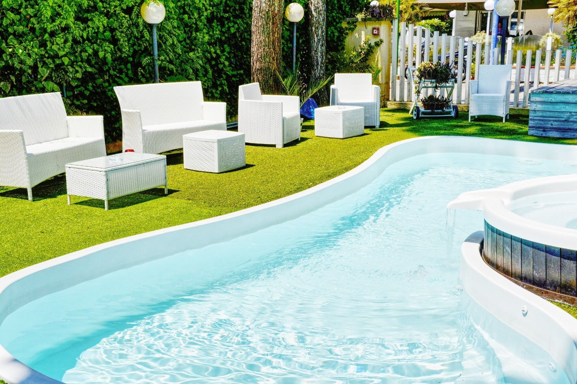 Una piscina con sillas de descanso blancas y una mesita, rodeada de un césped cuidado y una valla blanca.