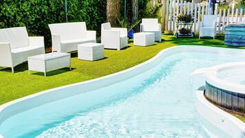 Una piscina con sillas de descanso blancas y una mesita, rodeada de un césped cuidado y una valla blanca.