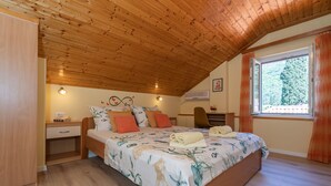 Quarto com teto de madeira, cama com roupa de cama floral, mesa de cabeceira, escrivaninha e janela com cortinas.