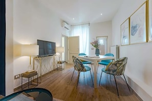 A modern hotel room with a flat-screen TV, a dining area with a table and chairs, and a large window with curtains.