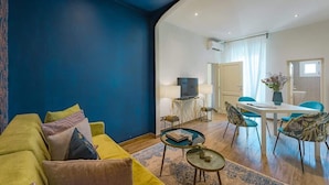A living room with a blue wall, a yellow sofa, a dining area with a table and chairs, and a television on a stand. - Macci A) Modern Elegant Apartment S. Ambrogio (Firenze)