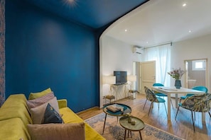 A living room with a blue wall, a yellow sofa, a dining area with a table and chairs, and a television on a stand.
