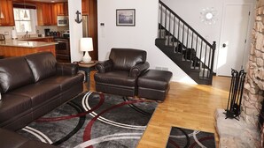 A living room with a brown leather sofa, a brown leather armchair, and a rug. There is a kitchen in the background with wooden cabinets and a refrigerator. A staircase is visible to the right.