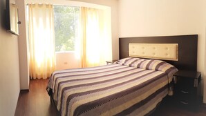 Une chambre à coucher avec un lit, une table de chevet, un téléviseur et une fenêtre avec des rideaux.