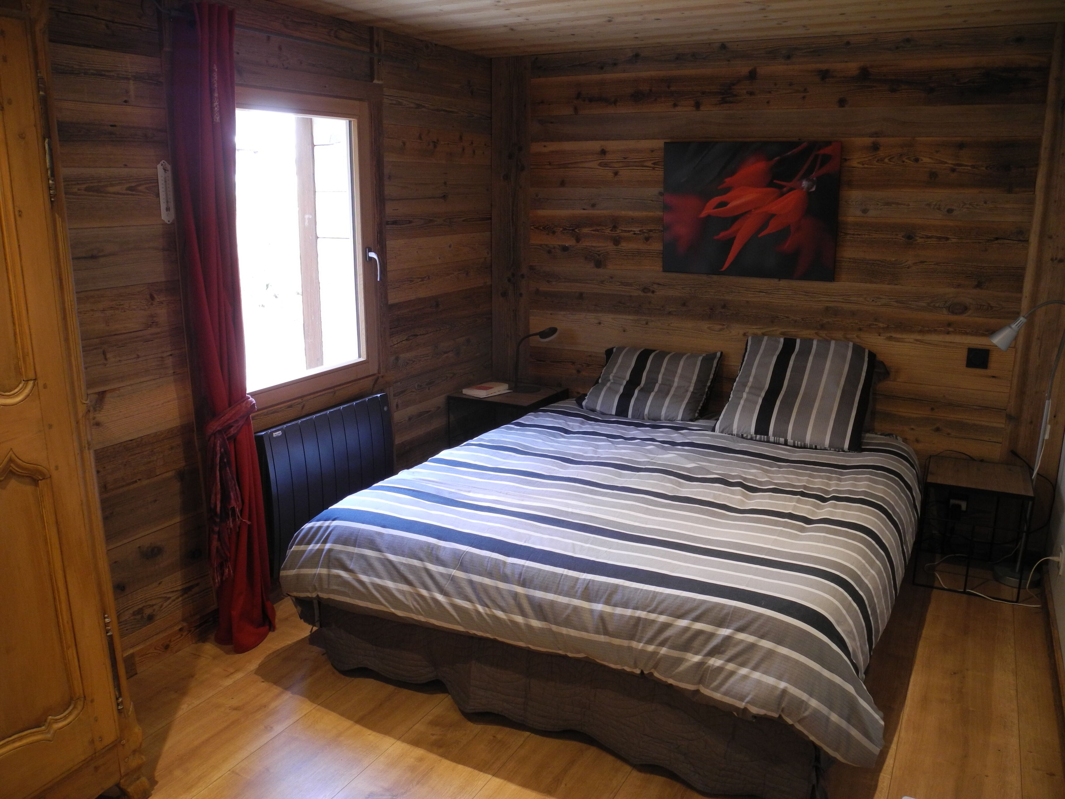 Een houten kamer met een bed, een nachtkastje, een raam met gordijnen en een schilderij aan de muur.