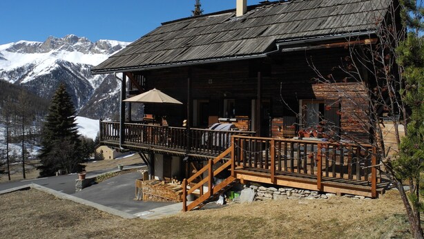 Uma cabana de madeira com varanda, chaminé e vista para montanhas cobertas de neve.
