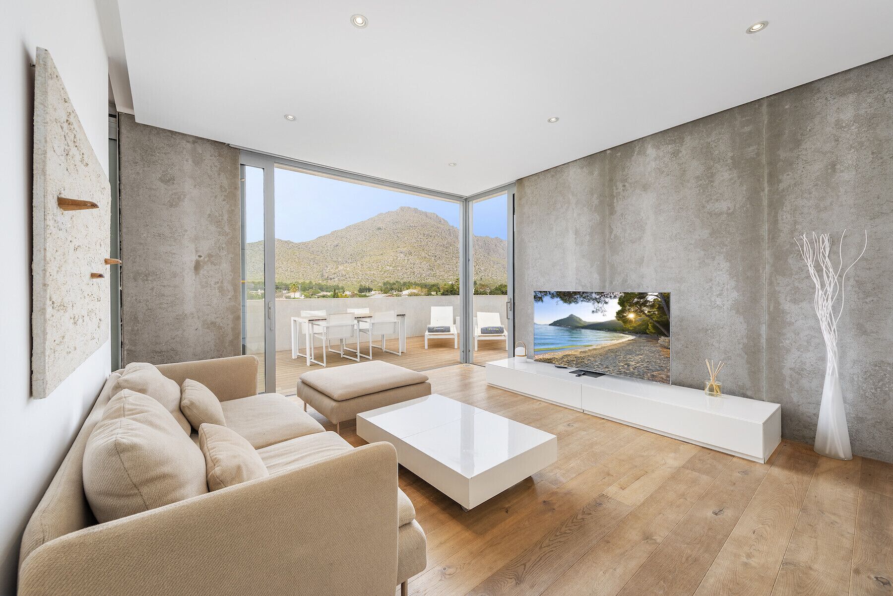 A modern living room with a large TV, beige sofas, a white coffee table, and a view of a mountainous landscape through large windows.