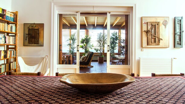 Una habitación con un recipiente de madera grande como centro de mesa, una estantería, una silla y vistas a un espacio exterior con plantas a través de una puerta de cristal.