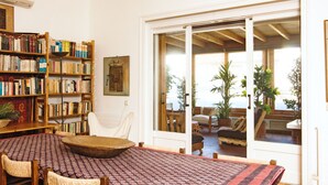 Una habitación con una gran estantería, una mesa de comedor con sillas y vistas a una zona de estar exterior con plantas a través de un gran ventanal corredero.