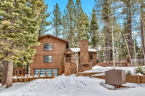 A two-story wooden cabin with a visible garage, surrounded by snow and trees.