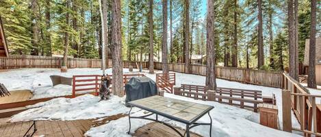 A wooden deck with a metal table, surrounded by a wooden fence and tall pine trees.