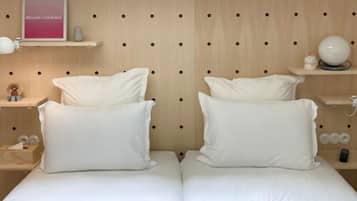 Una cama doble con ropa de cama y almohadas blancas, cabecera de madera con cortes circulares, estante pequeño con letrero rosa, espejo redondo y una figurita.
