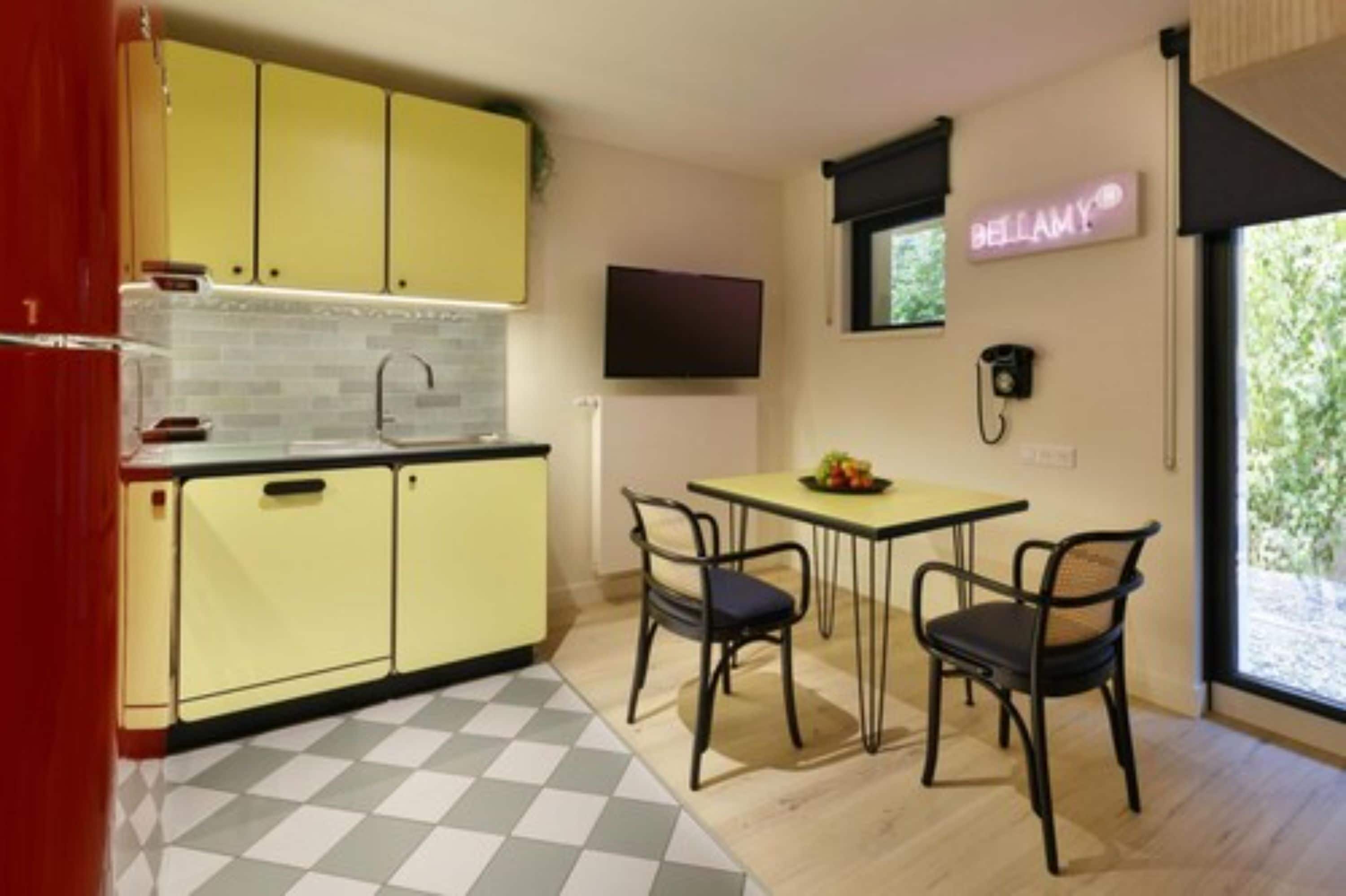 Een compacte keuken met gele kastjes, een geruit vloertje, een eettafel met twee stoelen, en een neonbord aan de muur.