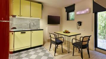 Een compacte keuken met gele kastjes, een geruit vloertje, een eettafel met twee stoelen, en een neonbord aan de muur.