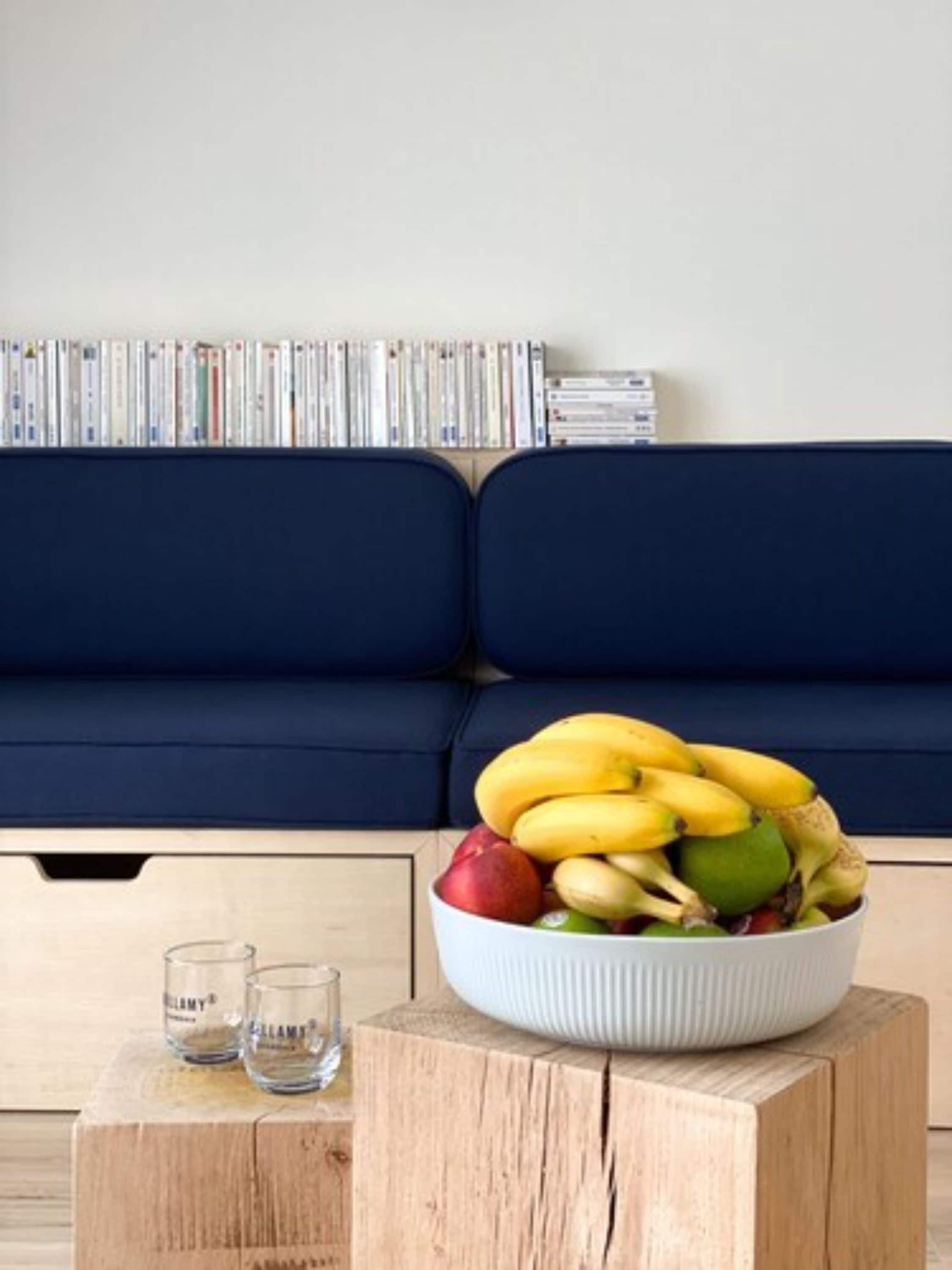 Una sala de estar con un sofá azul oscuro, una mesa de centro de madera con un frutero, y una fila de DVDs en la pared.