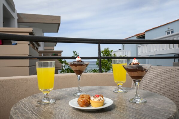 A table with two dessert glasses, a plate with pastries, and a glass of orange juice on a balcony overlooking a body of water. - Mare Blue & Suites (Apokoronas)