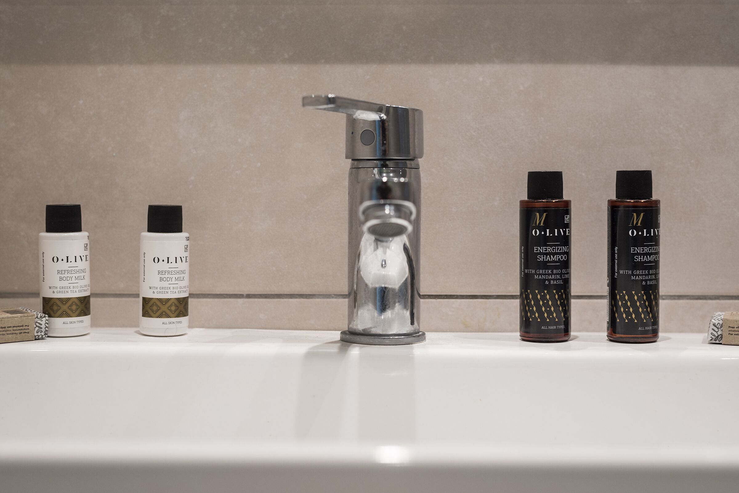 Un lavamanos de baño con un grifo moderno y cuatro botellas de leche corporal y champú O.L.I.V.E.