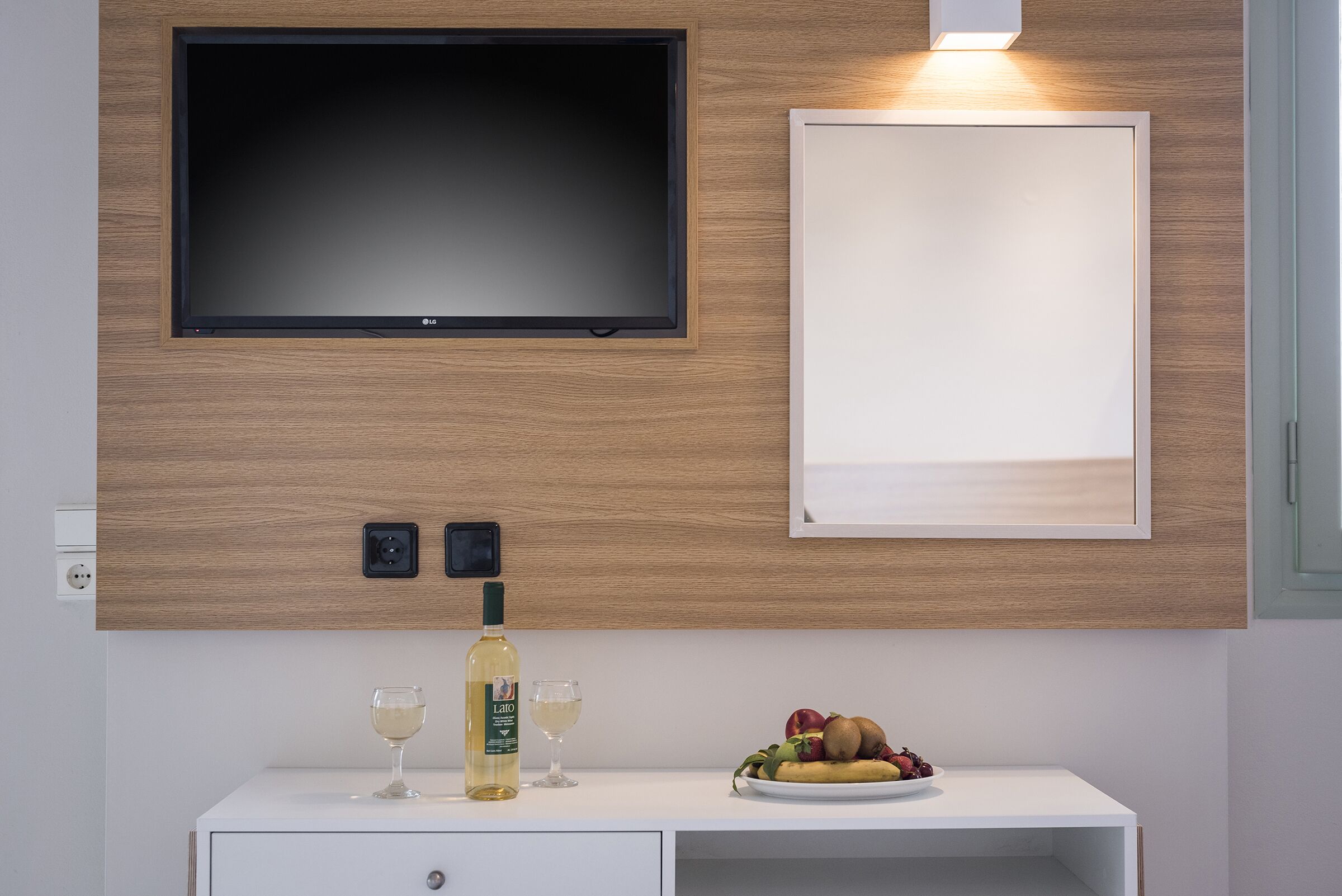 Una habitación moderna con televisión montada, un cuadro enmarcado, una mesita con una botella de vino, dos copas y un plato de fruta.