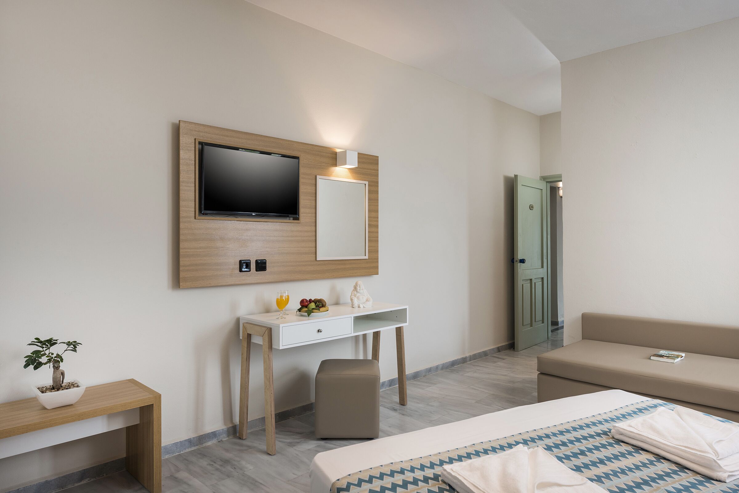 A modern hotel room with a bed, a desk with a flat-screen TV, a small plant, and a bench.
