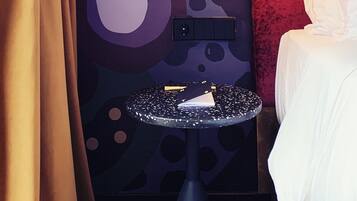 A hotel room with a patterned wall, a small round table with a book, a lamp, and a bed with white bedding.